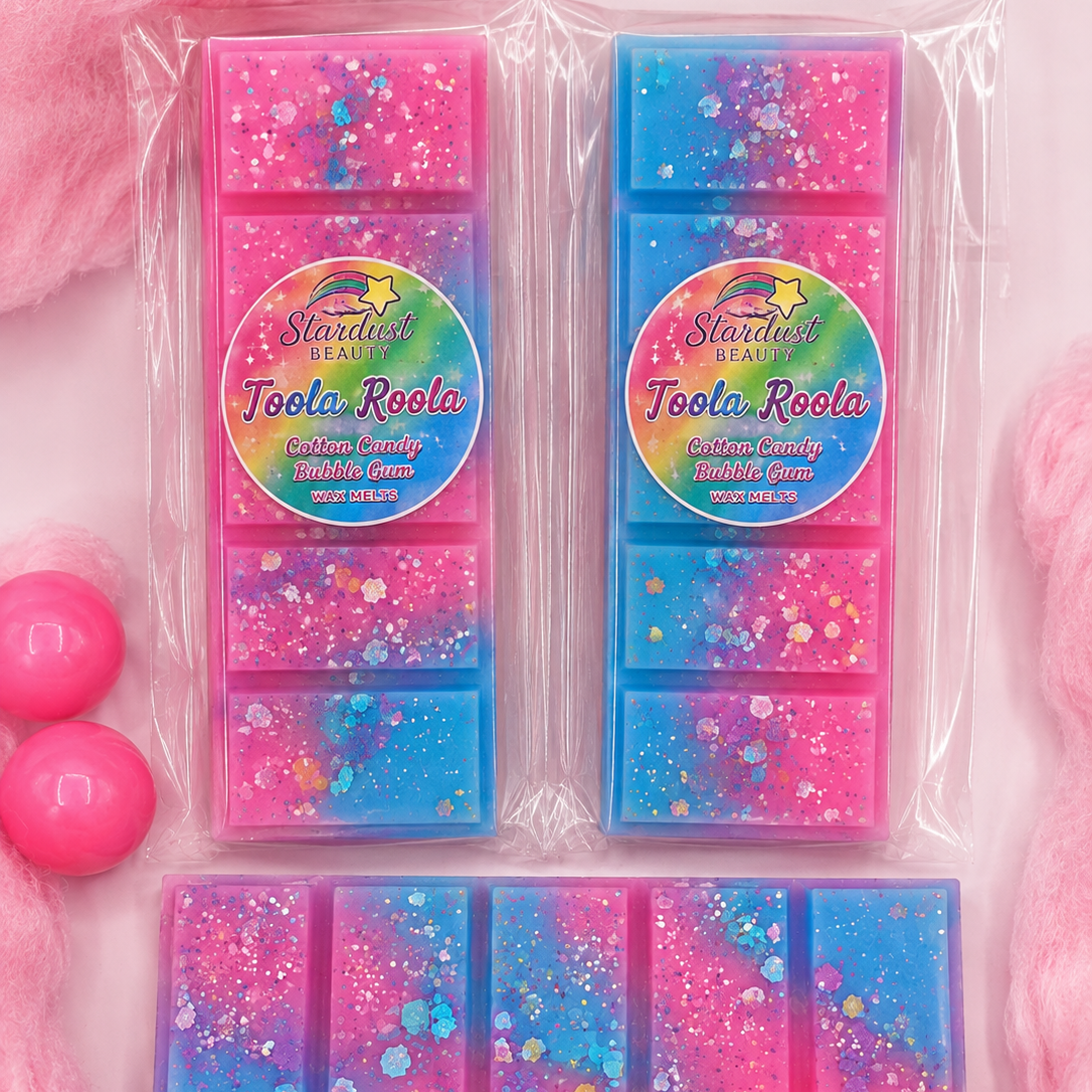 Three packaged rectangular coconut wax melt bars in glittery pink and blue, arranged on a pink background with fluffy pink fabric and pink balls.