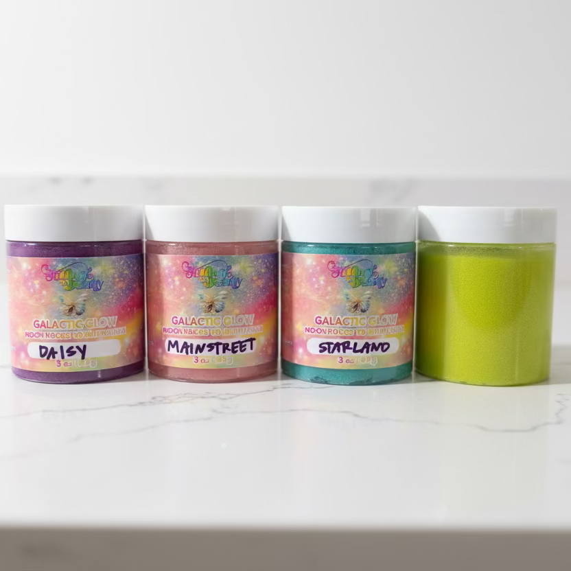 Four jars of Galactic Cosmetics glitter on a white surface in front of a black TV, with three labeled 'Daisy', 'Main Street', and 'Surfland' in pink, purple, and blue shades, and one lime green jar.